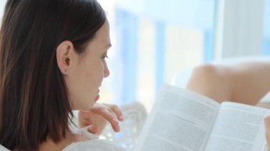 Güzel esmer kadın bir kitap okurken, beyaz koltukta ayakları masanın üzerinde, dikiz aynasından 4K video izliyor.