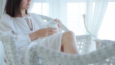 Beyaz retro-stil pijama giymiş esmer bir kadının portresi elinde bir fincanla koltukta oturur ve evdeki pencereden bakarken çay ya da kahve içer. Rahatlık ve stres yok, 4k