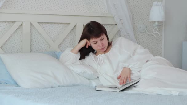 中世のショットの女性で白い夜のガウンはベッドルームでベッドの上にあり 朝の本を読み取りますとともに日光 4Kビデオ — ストック動画