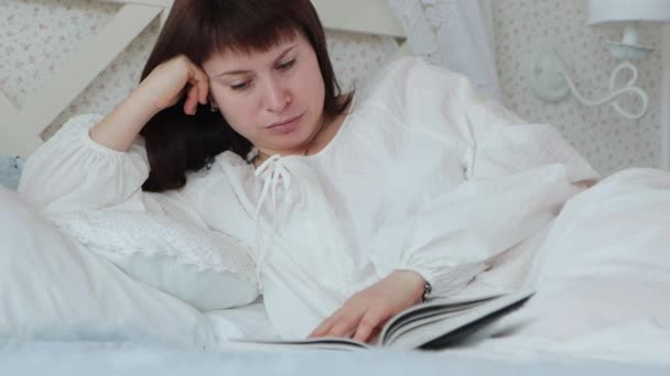 Μια Νεαρή Γυναίκα Ξαπλώνει Λευκό Νυχτικό Ένα Κρεβάτι Διαβάζει Βιβλίο — Αρχείο Βίντεο