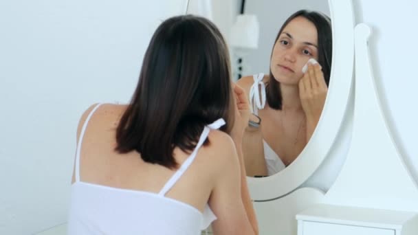 形象漂亮的年轻成年女子穿着白色睡衣坐在镜子前 用海绵在脸上喷粉 后视镜 化妆品概念的化妆 4K视频 — 图库视频影像