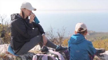 Dağın tepesinde çocuğu olan anne dinleniyor ve termostan çay içiyor, manzaraya hayran. Yürüyüş ve aktif yaşam tarzı kavramı.