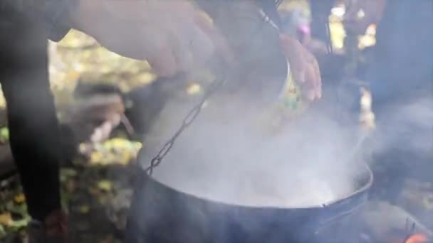 キャンプのキャンプファイヤーで釜で缶詰の肉でスープを調理する 手は鍋に缶詰の肉を注ぐ クローズアップ — ストック動画