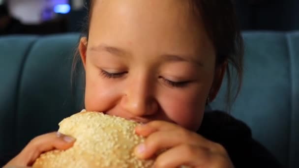 かわいい女の子がハンバーガーを食べる ハングリーベイビー ファーストフードカフェでハンバーガーを噛む クローズアップ — ストック動画