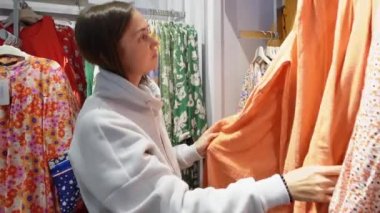 Genç bir kadın mağazadaki elbiseleri keşfediyor, şık seçimler yapıyor. Alışverişkolik alışveriş çılgınlığının tadını çıkarıyor, moda kıyafetleri heyecanlı seçiyor.