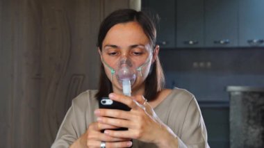 Maskeli bir kadın, nebulizör kullanıyor ve zatürree tedavisi sırasında akıllı telefona bakıyor..