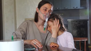 Anne ve kızı bir masada, maskeli bir kadın nebulizatörle ilaç soluyor. Anne zatürree tedavisi sırasında kızını nazikçe kucaklıyor ve sakinleştiriyor..