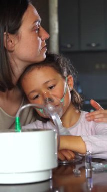 Bir anne ve kız oturuyor, sarılıyorlar. Anne, astım maskesi takan hasta bir çocuğa bakıyor. Üzgün kız, dokunaklı bir portrede annesine sarılıyor..