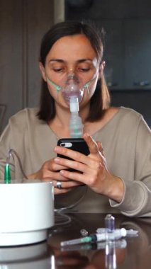 Maskeli bir kadın, nebulizörden nefes alıyor ve evde nefes alırken akıllı telefona bakıyor. Dikey görüntüler.