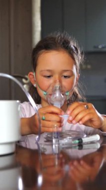 Evde tedavi gören küçük bir kız nebulizatörle nefes alıyor. Maske takıyor, solunum tedavisi görüyor. Dikey görüntüler.