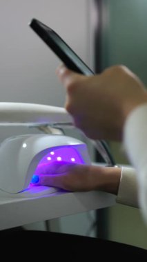 Kadın ultraviyole ışık altında jel cilası kuruturken kuaförde manikür yaparken akıllı telefon kullanıyor.