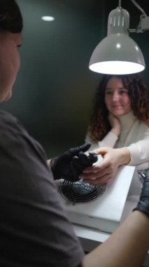 Manikürcünün tırnaklarını makineyle tedavi ederken genç bir kızın işlem sırasında duygusal olarak konuştuğu dikey video..