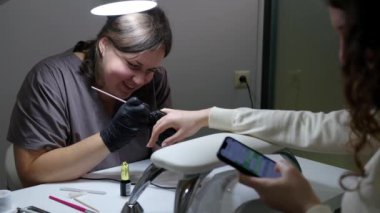 Müşteri için fırçayla tırnak boyarken gülümseyen profesyonel bir manikürcü. Genç kadın akıllı telefonuyla mesaj yazarken manikür yaptırıyor..