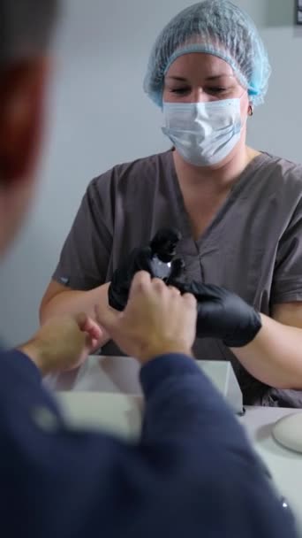 垂直录像拍摄了一位身穿制服 头戴头罩的美容师 为一位男性患者提供消毒手部治疗 注意手部清洁剂 — 图库视频影像