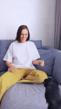 Genç kadın, küçük köpeği Mini Pinscher ya da Oyuncak Terrier ile kanepede oynaşmaktan hoşlanıyor. Kadın ve süs köpeği arasındaki komik etkileşimler. Dikey Video Biçimi.