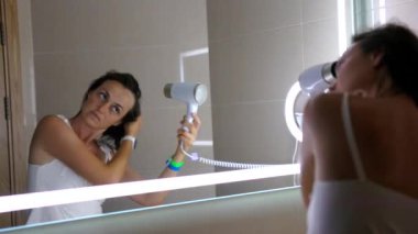 Banyoda genç bir kadın aynada kendine bakarken saçını saç kurutma makinesiyle kurutuyor. Kadın portresi, aynadaki yansıma..