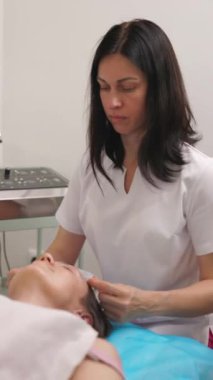 Bir güzellik uzmanı, müşterisini estetik ameliyatlara hazırlar. Müşterisi koltukta yatan hastaya cerrahi başlık takar. Dikey video biçimi.