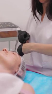 Kadın bir güzellik uzmanı lastik eldiven giyerken hasta bir kadın kanepede uzanıp estetik ameliyatlara hazırlanıyor. Dikey video biçimi.