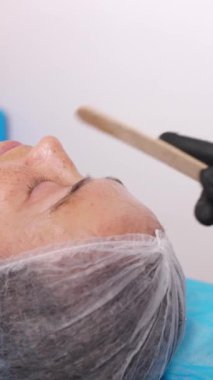 Bir kozmetik uzmanı profesyonel bir yüz bakımı sırasında yüzüne jel sürerken bir kadın gözlerini kanepeye kapatarak uzanıyor. Dikey video biçimi.