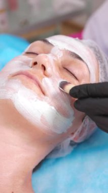 Spa tedavileri ve yüz bakımı. Profesyonel bir kozmetolojistin kadın müşterisinin yüzüne beyaz bir maske takarken çekilmiş dikey videosu..