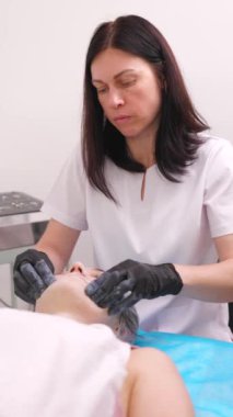 Kadın bir kozmetikçi hastanın yüzüne masaj yapıyor, nazikçe soymak, nemlendirmek ve spa tedavileri sırasında deriyi beslemek için makyaj yapıyor.