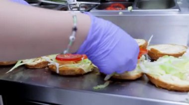 Hamburger yaparken lahana ve domates de dahil olmak üzere sebzeleri hamburger ekmeğine koyan bir fast food restoranında yakın plan bir aşçı. Fast food pişirmek için ideal..