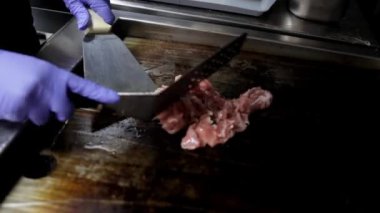 Bir restoran mutfağında, bir şefin elleri sobada spatula kullanarak doğranmış et kızartırken yakın planda gösterilir. Yemek pişirme teknikleri ve hazırlık süreci için mükemmel.
