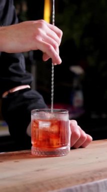 Bir barmenin elleri yakın planda kaşıkla alkollü bir içeceği buzla karıştırır. Dikey yavaş çekim video biçimi, kokteyl hazırlama ve içecek içeriği için mükemmel.