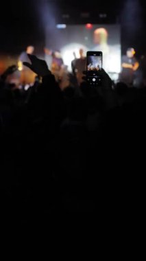 Sahnede bir orkestranın sahne aldığı bir konser sahnesinde seyirciler ayakta, dans ediyor ve arka planda ellerini sallıyorlar. İnsanların siluetleri enerjik bir atmosfer yaratır..