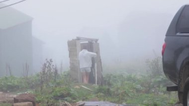 Bir kadın sisli kırsal bir ortamda eski, dökülen bir açık hava tuvaletine doğru yürüyor. Video, kırsal yaşamın kırsal cazibesini ve zorluklarını vurguluyor, harap olmuş yapıyı sergiliyor.