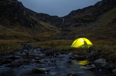 Alacakaranlıkta, Corrie Fee, Cairngorms Ulusal Parkı 'nda dağlarla çevrili bir nehrin yanındaki yeşil ışıklı kamp çadırının uzun pozlu görüntüsü.
