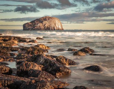İskoçya 'nın doğu kıyısındaki Bass Rock' ın uzun pozlu görüntüsü, kırılan dalgaların üzerinde güneş parlayan kayalık bir sahilden.