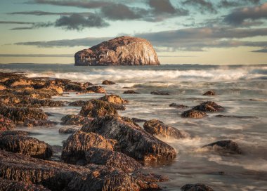 İskoçya 'nın doğu kıyısındaki Bass Rock' ın görüntüsü dalgaların üzerinde parlayan güneşli kayalık bir sahilden. Uzun pozlama