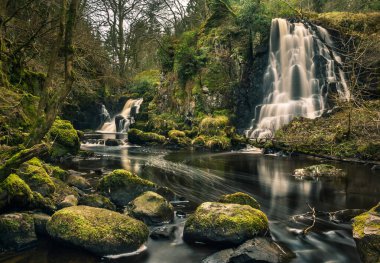 Long exposure shot of Linn Jaw Waterfalls, near Livingston, Scotland, with mossy rocks in the foreground and surrounding the waterfalls and white foam streaks in the water. West Lothian. UK clipart
