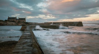 İskoçya 'nın Cove Limanı' ndaki taş duvara vuran dalgalar, bulutlara, denize ve arka plandaki yaşlı balıkçının evine akşam ışığı yansıyor.