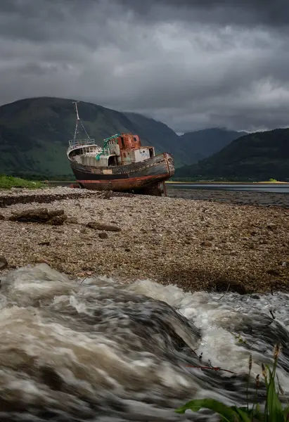 stock image Vertical landscape. Corpach shipwreck. Old fishing boat beached near Fort William. Highlands of Scotland. Running water in foreground. Clouds above Ben Nevis in background. Scotland. UK