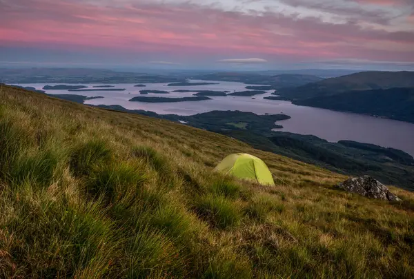 stock image Green tent wild camping on Ben Lomond at dusk with pink clouds, on the slope of the hill overlooking the lake, Loch Lomond and The Trossachs National Park, Scotland