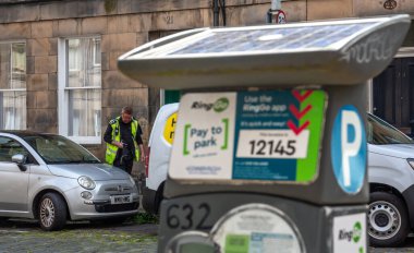 Edinburgh, İskoçya, 07.17.2024: Park görevlisi Edinburgh 'da park etmiş araçları ön planda parkmetreyle kontrol ediyor