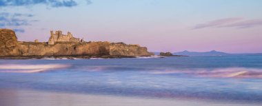 Ruins of Tantallon Castle from Seacliff beach, near North Berwick, Scotland, on a cliff by the sea at sunrise, with waves crashing on beach and pink sky reflected in the water clipart