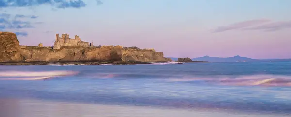 stock image Ruins of Tantallon Castle from Seacliff beach, near North Berwick, Scotland, on a cliff by the sea at sunrise, with waves crashing on beach and pink sky reflected in the water