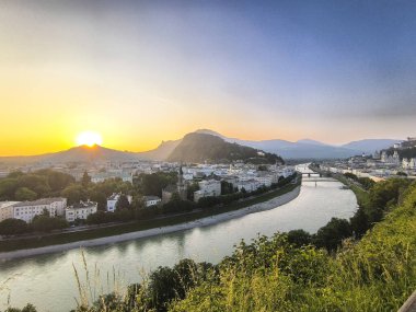 Gün doğumunda Monchsberg tepesinden Salzburg manzarası, Salzach nehri şehirden şehire ve tepelerin üzerinden yükselen güneş ve ön plandaki ağaçlar