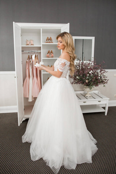 Свадьба. Невеста в красивом платье сидит на диване в помещении в белом интерьере студии, как дома. Трендовый свадебный стиль снят в полный рост. Молодая привлекательная брюнетка-кавказка, как невеста