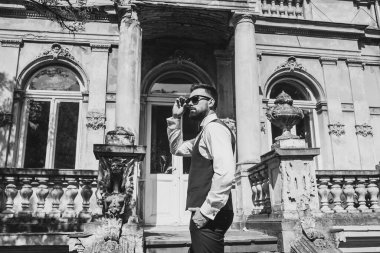 Açık mavi takım elbiseli yakışıklı Çinli genç bir adamın portresi ve beyaz atlet, güneşli bir günde modern şehir binalarıyla yürüyor ve uzaklara bakıyor, kendinden emin iş adamının yan görünüşü..