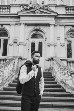 Açık mavi takım elbiseli yakışıklı Çinli genç bir adamın portresi ve beyaz atlet, güneşli bir günde modern şehir binalarıyla yürüyor ve uzaklara bakıyor, kendinden emin iş adamının yan görünüşü..