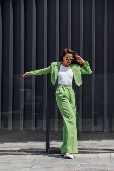 Fashionable elegant confident woman wearing trendy pink sunglasses, silk headscarf, suit blazer, with green bag, posing in street of European city. Outdoor fashion portrait. Copy, empty space for text