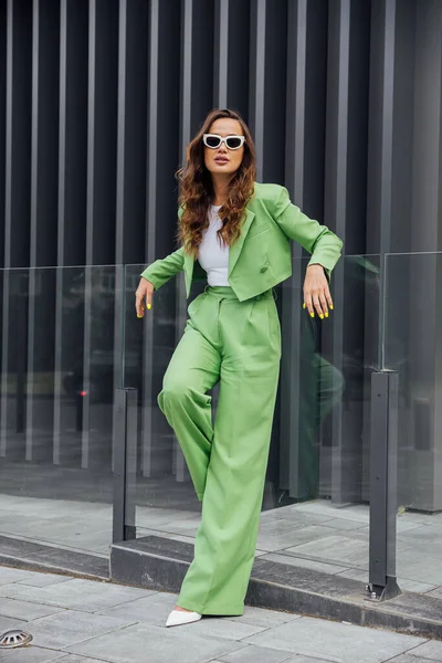 Fashionable elegant confident woman wearing trendy pink sunglasses, silk headscarf, suit blazer, with green bag, posing in street of European city. Outdoor fashion portrait. Copy, empty space for text
