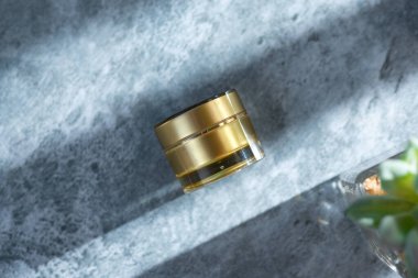 Gri yüzeyde altın renkli krema kavanozu. Gölge kaplamalı. Güzellik ürünleri için model.