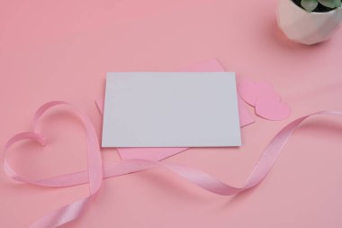 Boş bir kart pembe bir zarfın içinde, iki kağıt kalbin ve kalp şeklinde pembe bir kurdelenin yanında duruyor. Kart modeli