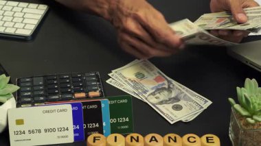 FINANCE etiketli birkaç tahta blok ve siyah hesap makinesinin önünde birkaç kredi kartı bulunan bir masanın üzerindeki dolar banknotlarını sayan bir elin görüntüsü..