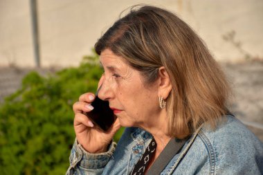 Yaşlı kadın cep telefonuyla iletişim kuruyor, endişeli görünüyor. Odaklanmış bakışı önemli ya da ilgili bir konuşma olduğunu gösteriyor..